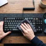 Person cleaning a mechanical keyboard with gel and a cloth on a wooden desk.